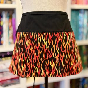 May include: A black waist apron with a flame pattern. The apron features a solid black top section and a lower section with a repeating design of red, orange, and yellow flames on a black background. The apron is displayed on a mannequin.