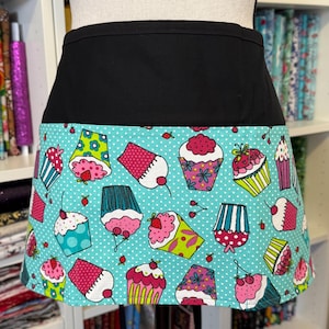 May include: A black waist apron with a turquoise bottom featuring a pattern of colorful cupcakes. The cupcakes have pink, red, yellow, and green frosting and are decorated with cherries and sprinkles. The apron is displayed on a mannequin.