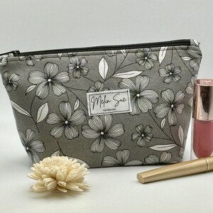 May include: A gray floral print cosmetic bag with a black zipper, a white label that says "Melin Sue Handmade", a pink lip gloss, and a gold mascara tube. A white flower is in the foreground.