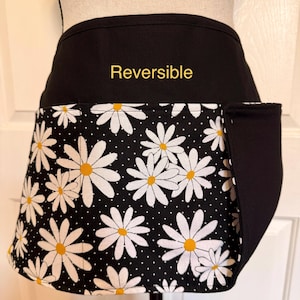 May include: Reversible black and white daisy print apron with a pocket. The apron features a black top and a daisy pattern on a black background with white polka dots. The word "Reversible" is printed in gold.