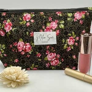 May include: A floral print cosmetic bag with a black zipper, featuring pink roses and green leaves on a black background. A tube of pink lipstick and a gold-colored mascara tube are next to the bag. A decorative cream-colored flower is in front.