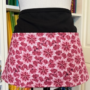 May include: A black waist apron with a floral pattern. The apron's top is solid black, while the lower portion features a repeating design of pink and white flowers. The flowers have multiple layers of petals.