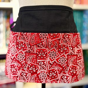 May include: A half apron with a black waistband and a red bandana print skirt. The skirt features a repeating pattern of white paisley and floral designs on a red background. The apron is displayed on a mannequin.
