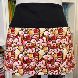 May include: A black waist apron with a colorful pattern of desserts. The pattern includes ice cream cones, cupcakes, donuts, and macarons in shades of pink, red, yellow, and brown. The apron is designed for kitchen or craft use.