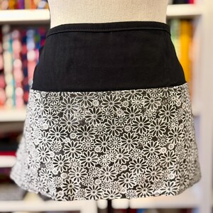 May include: A black and white daisy print apron. The apron has a solid black waistband and a skirt section covered in a pattern of white daisies on a black background. The apron is displayed on a mannequin.
