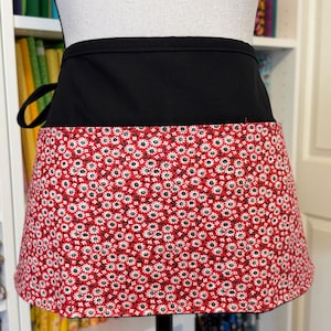 May include: A black and red half apron. The top portion is solid black, while the bottom features a red floral pattern with white and black accents. The apron is tied at the waist with black ties.