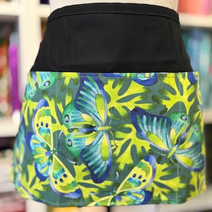 May include: A black waist apron with a vibrant butterfly pattern. The apron features a solid black waistband and a lower section adorned with large butterflies in shades of blue, green, and yellow against a green background.