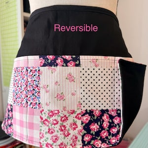 May include: Reversible black and patchwork apron. The patchwork features pink floral, polka dot, and gingham patterns. The apron has a black pocket and the word "Reversible" in pink.