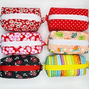 May include: Six small, rectangular fabric pouches with zippers. The pouches feature various floral and patterned designs in red, pink, black, and yellow. Each pouch has a white or red zipper and a small metal loop on the side.