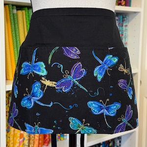 May include: Black waist apron with a pocket featuring a colorful dragonfly pattern. The dragonflies are in shades of blue, purple, and gold, set against a black background. The apron is designed for kitchen or craft use.