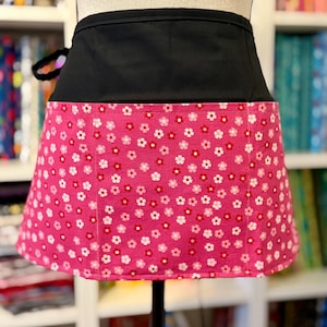 May include: A black waist apron with a pink floral patterned skirt. The skirt features a repeating pattern of small white and red flowers on a bright pink background. The apron is designed for use in a kitchen or craft setting.