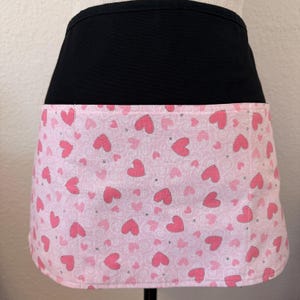 May include: A black waist apron with a pink heart-patterned pocket. The pocket fabric features various sizes of pink hearts with silver accents on a light pink background. The apron is designed to be worn around the waist.