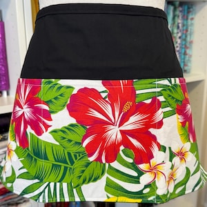 May include: A black waist apron with a vibrant floral design. The apron features a white background with large red hibiscus flowers, green leaves, and smaller white and yellow flowers. The top portion is solid black.