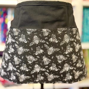May include: A black waist apron with a patterned lower section. The pattern features detailed illustrations of bees in shades of gray and white against a black background. The upper section of the apron is solid black.