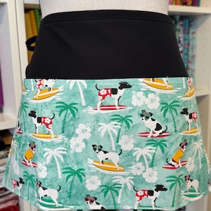 May include: A black waist apron with a tropical print featuring dogs surfing on yellow and red surfboards, palm trees, and white flowers against a teal background. The apron is tied at the waist.
