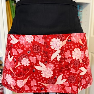 May include: A black waist apron with a vibrant red floral print. The apron features a bold pattern of pink and white flowers, with a black waistband. The apron is designed for kitchen or craft use.