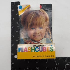 May include: A Sylvania Blue Dot Flashcubes package featuring a photograph of a child with blonde hair. The packaging text reads Sylvania Blue Dot Flashcubes 3 Cubes/12 Flashes. A black ruler is visible along the side, showing a height of 7 inches.