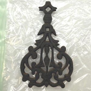 May include: A dark brown cast iron trivet featuring an elongated, symmetrical design with intricate scrollwork, leaf-like motifs, and a central vertical stem that culminates in a pointed top section with a small diamond-shaped cutout.