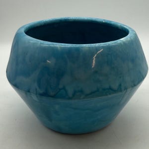 May include: A wide, bowl-shaped ceramic planter featuring a glossy, variegated glaze in shades of bright turquoise and deep ocean blue. The vessel has a smooth, rounded rim and a tapered base, showcasing a swirling, marbled color pattern.