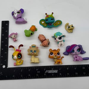 May include: A collection of ten small plastic animal figurines, including a purple rabbit, a green turtle, a butterfly, a tan cat, a white lamb, a yellow rabbit, a tan dog, a tiger, and a purple dog, arranged next to an L-shaped ruler.