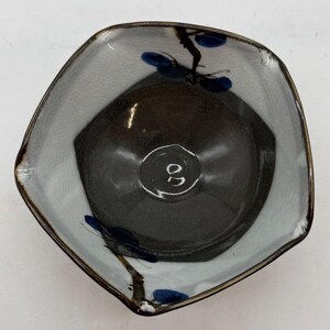 May include: A small, irregularly shaped ceramic bowl featuring a dark brown, smooth interior and a light gray, crackled glaze exterior. The rim is decorated with hand-painted dark blue floral motifs and thin brown stems, creating a contrast.