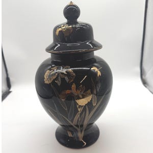 May include: A black ceramic ginger jar with a matching lid featuring a pointed finial. The jar is decorated with gold and silver metallic illustrations of iris flowers and leaves, with a small dragonfly depicted on the upper right side.