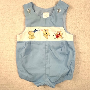 Op de afbeelding: Een lichtblauwe mouwloze romper met een crèmekleurig borstpaneel met geborduurde beige teddyberen die muziekinstrumenten bespelen. Het kledingstuk heeft twee doorzichtige knopen op de schouders en een geplooid voorpand met een ronde halslijn.