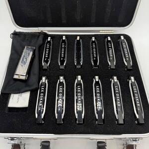 May include: A collection of twelve Lee Oskar harmonicas arranged in a silver-toned carrying case with black foam inserts. Each harmonica features a black and silver body with lettered labels. One harmonica is stored in a black fabric pouch.