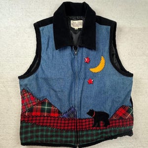 Puede incluir: Un chaleco de mezclilla azul con cuello negro y cierre de cremallera frontal. La parte inferior tiene un diseño de patchwork de tela escocesa roja y verde. Los apliques incluyen una luna creciente amarilla, dos estrellas rojas y un oso negro.