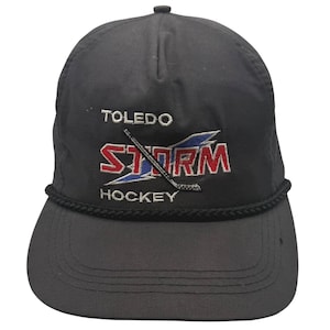 May include: A black baseball cap featuring an embroidered logo for the Toledo Storm hockey team. The design includes the text TOLEDO STORM HOCKEY in white and red lettering with a blue lightning bolt graphic and a black hockey stick across the front.