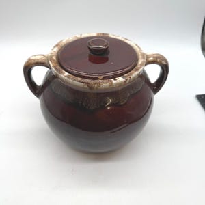 May include: A round, dark brown ceramic sugar bowl with two side handles and a matching lid. The rim of the lid and the upper shoulder of the bowl feature a decorative drip glaze effect in shades of cream and light brown, contrasting with the glossy, solid dark brown finish of the main body.