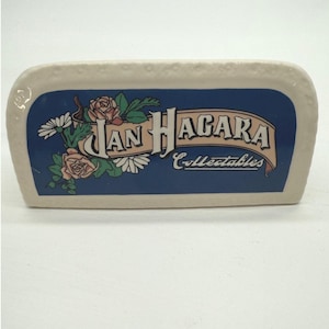 Könnte beinhalten: Ein rechteckiges Keramik-Schmucktablett mit cremefarbenem Rand und blauem Mittelteil. Die Mitte zeigt den Text "Jan Hagara Collectibles" mit floralen Akzenten aus rosa Rosen und weißen Gänseblümchen.