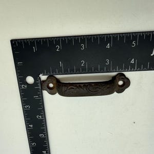 Könnte beinhalten: Ein dunkelbrauner Schubladengriff aus Gussmetall mit einem verzierten, geprägten Blumenmuster. Der Griff hat auf beiden Seiten zwei kreisförmige Befestigungslöcher. Ein schwarzes L-förmiges Metalllineal liegt oben links und zeigt 10 cm an.