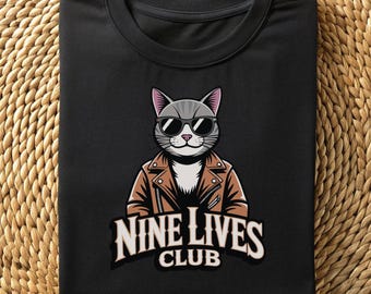 May include: A black crew neck t-shirt featuring a graphic of a gray cat wearing black sunglasses and a brown leather jacket. Below the cat, the text reads NINE LIVES CLUB in a bold, white, arched font. The design is centered on the chest area.