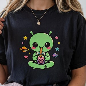 Può includere: T-shirt nero con un alieno verde dei cartoni animati che beve bubble tea. L'alieno è circondato da stelle e pianeti. Il design è colorato e stravagante, attraente per i fan dello spazio e dell'estetica kawaii.