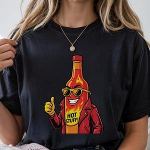 May include: Black t-shirt featuring a cartoon hot sauce bottle character wearing sunglasses and a red jacket, giving a thumbs-up. The text "HOT STUFF!" is on the bottle. The shirt is a comfortable fit.