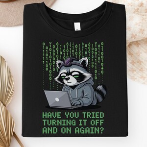 May include: A black t-shirt featuring a graphic of a raccoon wearing a grey hoodie and a headset while using a laptop. Behind the raccoon is a vertical pattern of green binary code. The text below reads, HAVE YOU TRIED TURNING IT OFF AND ON AGAIN?