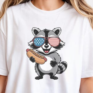 Può includere: Una t-shirt bianca a maniche corte con un'illustrazione grafica di un procione grigio e bianco che indossa occhiali da sole con il motivo della bandiera americana rossa, bianca e blu. Il procione sorride e tiene un hot dog.