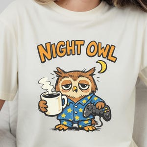 May include: Cream t-shirt with a cartoon owl in blue star-patterned pyjamas, holding a coffee mug and a games controller. The text "NIGHT OWL" is above the owl in orange and black. A crescent moon is also present.