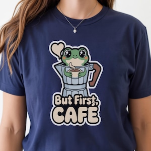 May include: Navy blue t-shirt featuring a cartoon frog holding a coffee cup inside a silver coffee maker. The text "But First, CAFE" is printed below the frog in a white and beige font. The shirt is made of a soft material.