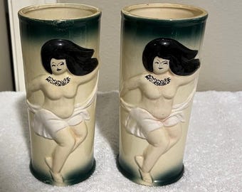 Pode incluir: Par de copos tiki vintage em cerâmica. Cada copo apresenta um relevo de uma mulher dançando com cabelo preto e saia branca. Os copos têm um corpo creme com borda e base verde escuro.