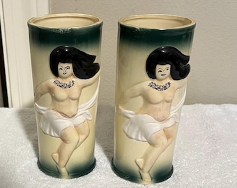 Pode incluir: Duas canecas tiki de cerâmica vintage. Cada caneca apresenta um relevo de uma mulher dançando com cabelo preto, uma saia branca e um colar de flores. As canecas têm um corpo creme com a parte superior e a base verdes.