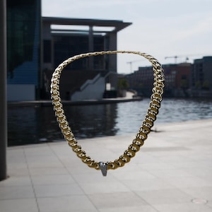 May include: A gold-toned cuban link chain necklace featuring a single, small, silver-toned triangular pendant at the center. The chain consists of interlocking, rounded metal links with a polished, reflective finish that catches the light.