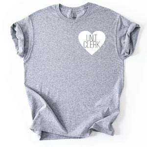 Peut inclure: Un t-shirt gris chiné avec un motif en forme de cœur blanc. Le cœur contient les mots "UNIT CLERK" dans une police simple sans empattement. Le t-shirt a des manches retroussées et un col rond.