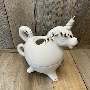 Puede incluir: Una cremera o jarra pequeña de cerámica blanca con forma de unicornio, con detalles dorados en el cuerno, la melena y el asa. El unicornio tiene los ojos cerrados y está sobre una superficie de madera.