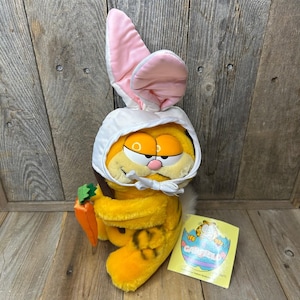 Puede incluir: Un peluche de Garfield disfrazado de conejito de Pascua. El gato amarillo lleva un gorro blanco con orejas forradas de rosa y sostiene una zanahoria. Se ve una etiqueta con el logotipo de Garfield. El juguete está sobre una superficie de madera.