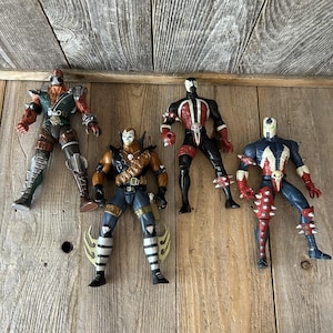 May include: Four action figures of Spawn characters. The figures are in various poses and feature detailed designs with a mix of colors including red, black, blue, and brown. They are set against a wooden background.