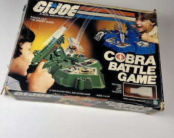 Puede incluir: Una caja vintage del juego Cobra Battle Game de G.I. Joe. La caja presenta ilustraciones de niños jugando, un lanzamisiles verde y el título del juego. La caja está desgastada, con el texto "A Real American Hero".