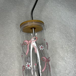 May include: A clear glass tumbler with a bamboo lid and a stainless steel straw. The tumbler is decorated with pink bows and white daisies. The straw is curved. The tumbler is designed for cold beverages.