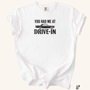 Peut inclure: Un t-shirt blanc à manches courtes avec une impression graphique noire sur la poitrine. Le motif comprend le texte "YOU HAD ME AT" au-dessus d'une silhouette noire de voiture ancienne, avec le mot "DRIVE-IN" imprimé en lettres noires grasses.